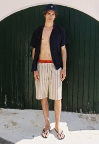 Marineblauw kortshirt met korte mouwen, gestreepte beige shorts met een rode tailleband, slippers, staand voor een groene houten deur.