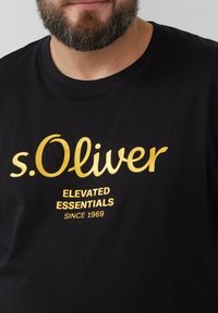 Schwarzes Baumwoll-T-Shirt mit einem goldenen Logo, das "s.Oliver" zeigt, sowie dem Text "ELEVATED ESSENTIALS" und "SINCE 1969" in kleinerer Schrift.