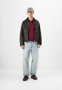 Jeune homme portant une casquette grise, une veste en cuir brun foncé, un polo bordeaux, un jean clair à jambes larges, et des chaussures brun foncé, se tenant contre un fond blanc.