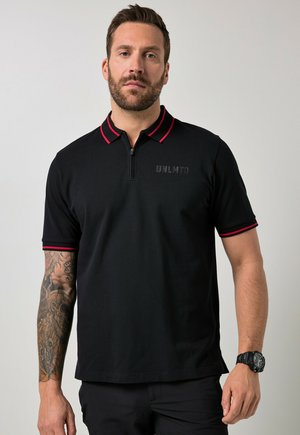 Uomo con barba che indossa una polo nera con rifiniture rosse e la scritta "UNLMTD", mostra una manica tatuata completa sul braccio sinistro e un orologio da polso nero.