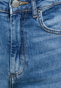 Zbliżenie przedniej części niebieskich dżinsów z denimu z metalowym guzikiem, szlufką na pasek, przeszyciami i szczegółami kieszeni.