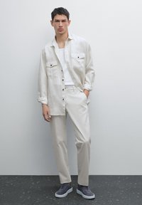 Light beige linen shirt with two chest pockets, paired with cream trousers and dark gray sneakers. Smooth texture and relaxed fit.