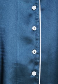 Satinbluse in Dunkelblau mit glatter Textur, ausgestattet mit weißen Paspeln entlang der Knopfleiste und sechs weißen Knöpfen an der Vorderseite.