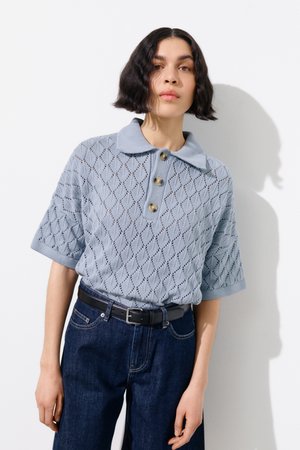 Persoon met kort donker haar draagt een lichtblauw gebreid poloshirt met knopen en donkere spijkerbroek met een zwarte riem.