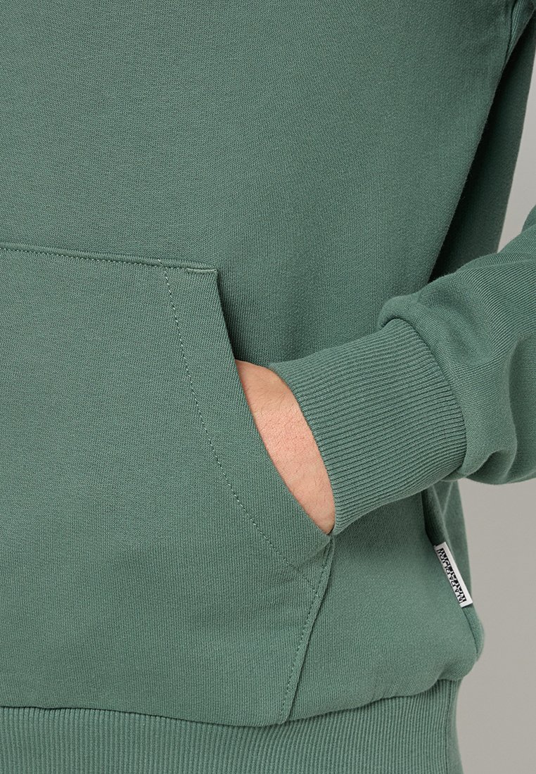 Grüner Pullover mit gerippten Bündchen und Tasche; weiche Textur; verfügt über ein kleines Logotag an der Seite. Nahaufnahme einer Hand in der Tasche.