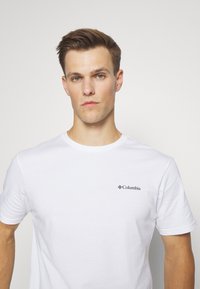 T-shirt en coton blanc avec un col rond, des manches courtes et un logo Columbia noir sur la poitrine gauche. Texture lisse, coupe décontractée.