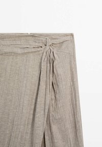 Tissu beige clair avec texture côtelée verticale noué en haut, montrant un gros plan de la zone de la ceinture d’un vêtement.