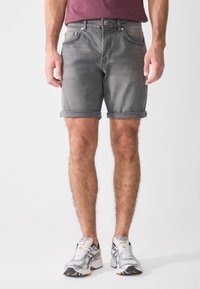 Shorts en denim gris avec un ourlet roulé, cinq poches, et une fermeture à boutons et zip. Portés avec des chaussures de sport blanches et grises.