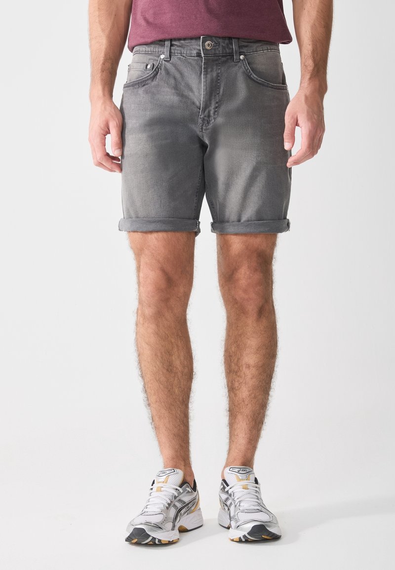 Shorts en denim gris avec un ourlet roulé, cinq poches, et une fermeture à boutons et zip. Portés avec des chaussures de sport blanches et grises.
