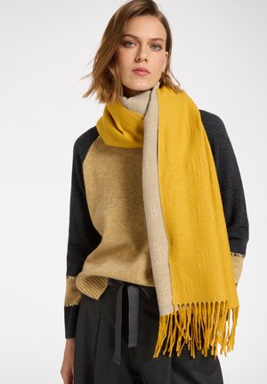 Vrouw draagt een tweekleurige gebreide trui, een geel-beige sjaal met franjes om haar nek gewikkeld, en donkere broek met een strik aan de voorkant.