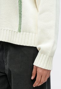 Pleteni kremasti džemper s rebrastim rubom i manžetama, s ukrasnim zelenim šavom sa strane. Nošen preko tamnih kord hlača.