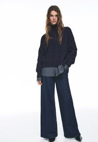 Marineblauer Oversize-Pullover, kombiniert mit einem blau-weiß karierten Hemd, weiten dunklen Jeanshosen und schwarzen spitzen Schuhen.