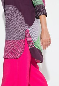 Top multicolore con motivi astratti in viola, rosa e verde; maniche corte arrotolate; abbinato a pantaloni a gamba larga rosa brillante.