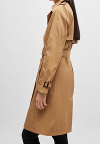 Widok z boku osoby w beżowym płaszczu do kolan z paskiem i detalami klamry, noszącej czarne spodnie, na jednolitym tle.