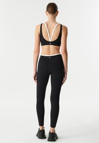 Black athletic crop top with white accent straps, V-back design; paired with black leggings. Model shown from the back, wearing black sneakers.