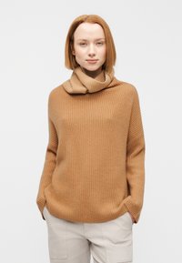 Καφέ ζιβάγκο με ραβδώσεις και διπλωμένο γιακά, με άνετη εφαρμογή και πλαϊνές τσέπες, συνδυασμένο με ανοιχτό μπεζ παντελόνι.