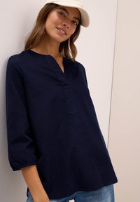 Vrouw in een marineblauwe langemouwtop met elastische manchetten, lichte spijkerbroek en een beige emmerhoed, glimlachend leunend tegen een muur.