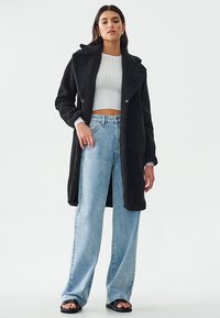 Abrigo largo de piel sintética negra con un amplio collar, combinado con un suéter corto blanco y unos vaqueros de tiro alto de color azul claro. Sandalias negras de tacón abierto.