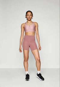 Sujetador deportivo rosa claro con tirantes ajustables y cierre de presión frontal, combinado con pantalones cortos de cintura alta a juego. Lleva zapatillas negras.