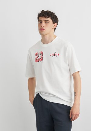 Young man with curly hair wearing a white t-shirt with red "23" and Jordan logo, and dark pants, standing against a plain background.