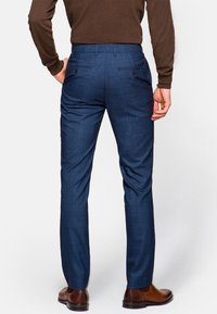 Granatowe spodnie w kratę o dopasowanym kroju, z dwiema tylkami kieszeniami z zapięciami na guziki oraz gładkim, teksturowanym materiałem. Widoczne brązowe skórzane buty.