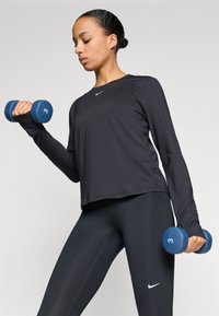 Μαύρη αθλητική μπλούζα με μακριά μανίκια και στενή εφαρμογή, συνδυασμένη με μαύρες κολάν. Μπλε αλτήρες με την ένδειξη "3" κρατούνται και στα δύο χέρια.