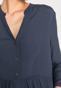 Kvinna iklädd en mörkblå texturerad blus med V-ringning och tre knappar fram, som visar en del av halsen och axeln.