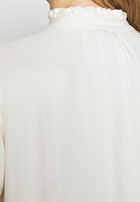 Weiße Bluse mit rüschigem Stehkragen, weichem Griff und Falten auf der Rückseite. Glattes Gewebe mit schimmerndem Finish.