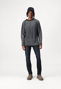 Grau und schwarz gestreifter Strickpullover mit weitem Ausschnitt, kombiniert mit dunklen Jeans und beigen Schnürschuhen. Schwarze Kappe mit Logoakzent.