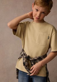 Beige bomulls-t-shirt med bröstficka, bärs med en knuten svart och grå rutiga flanellskjorta runt midjan, ihop med blå jeans.