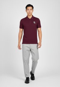 Burgundské polo tričko s krátkými rukávy, má skládací límec a malé vyšívané logo. Nosené s světle šedými kalhotami a černými botami.