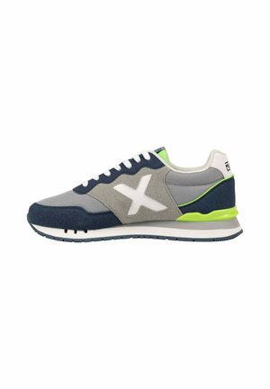 Zapatilla gris y azul marino con cordones blancos, logotipo blanco en forma de "X" y detalles en verde neón en la suela y la lengüeta del talón, vista desde el lado exterior.