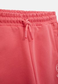 Pantaloni della tuta rosa con vita elastica e cordino, texture liscia e design minimalista. Include un piccolo logo dettagliato sul lato.