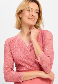 Rosa Spitzenoberteil mit Pailletten, gewelltem Ausschnitt und dreiviertel Ärmeln. Texturiertes Blumenmuster verleiht Detail. Glattes, tailliertes Design darunter.