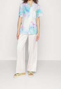 Camicia a maniche corte con bottoni in un motivo tie-dye pastello nei toni di blu, rosa e giallo; abbinata a pantaloni larghi rosa chiaro e sandali verdi.