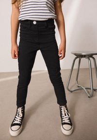 Jeans ajustados negros con diseño de cinco bolsillos y cierre de botón. Combinados con zapatillas altas negras con suelas y cordones blancos.