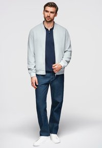 Un giovane uomo è in piedi indossando una giacca leggera grigia con zip sopra una maglietta polo blu navy, jeans blu e scarpe da ginnastica bianche su uno sfondo bianco uniforme.