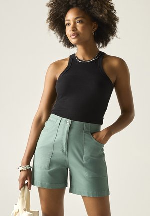Mujer con cabello rizado que lleva una camiseta sin mangas negra y pantalones cortos verdes, sosteniendo un bolso de mano crema con una mano, de pie frente a un fondo liso.