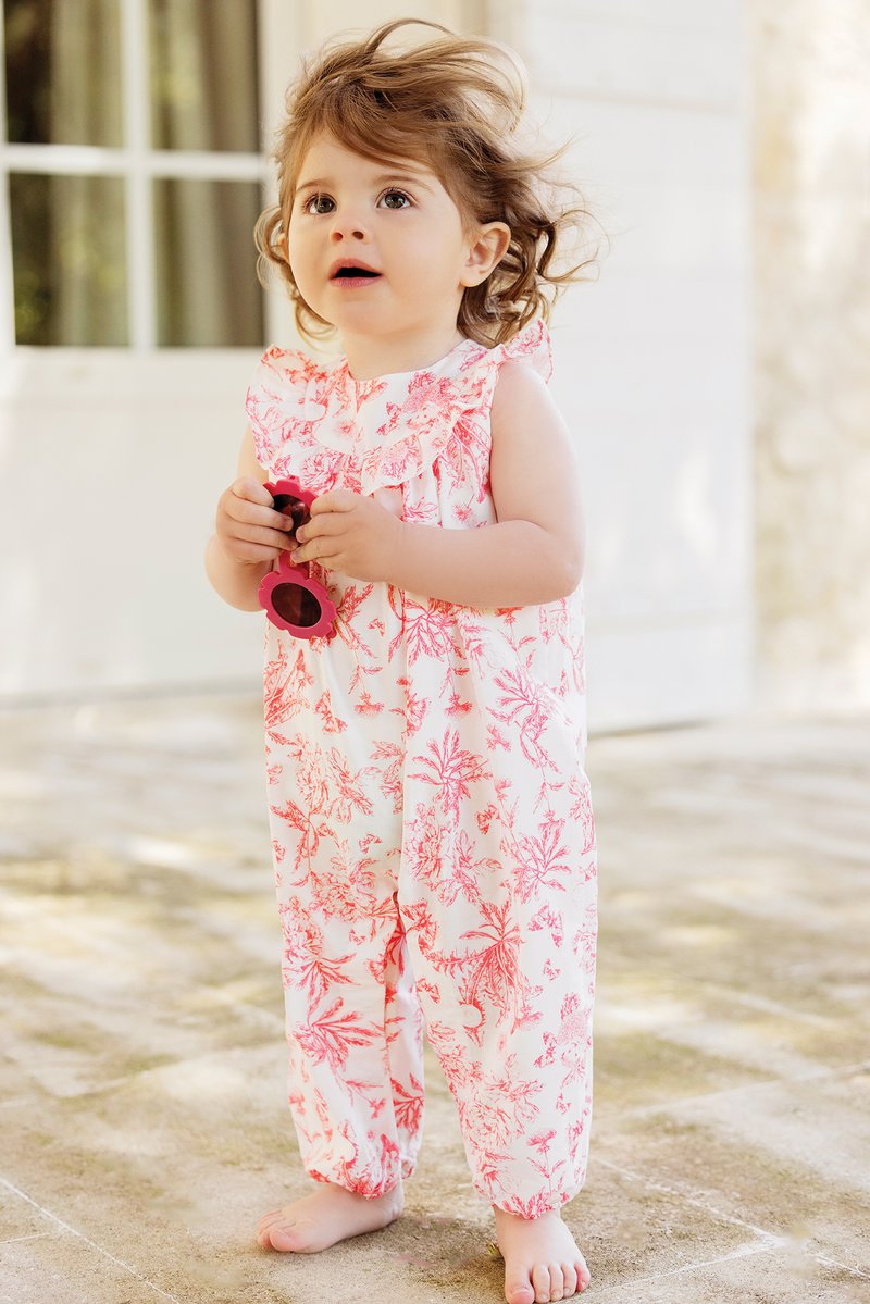 Macacão branco com padrão floral rosa, com ombros franzidos. A criança segura óculos de sol rosa, em pé sobre uma superfície de pedra clara.