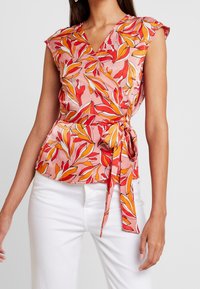 Blomstret pink top med korte ærmer, der har røde og gule bladmønstre, en V-udskæring og et bånd i livet for en tætsiddende silhuet.