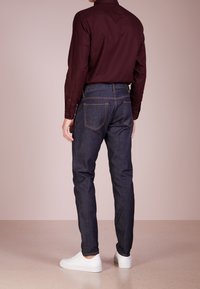 Vaqueros de mezclilla oscura con corte ajustado, costuras naranjas contrastantes y bolsillos traseros. Combinados con una camisa de manga larga en un burdeos profundo y zapatillas blancas.