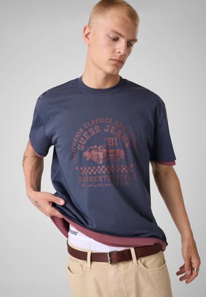 Młody mężczyzna z krótkimi włosami, ubrany w granatowy T-shirt z grafiką, beżowe spodnie i brązowy pasek, poprawiający koszulę jedną ręką.
