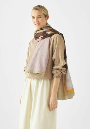 Vrouw glimlachend, draagt een beige blouse met opgerolde mouwen, een crèmekleurige rok en een grote, meerkleurige sjaal over de schouders gedrapeerd.