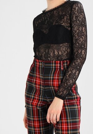 Osoba ubrana w czarny przezroczysty koronkowy top z długim rękawem na czarnym staniku bralette, współgrający z czerwono-czarnymi spodniami w kratę z wysokim stanem.