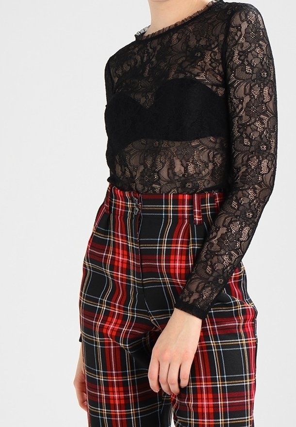 Personne portant un haut en dentelle noire transparente à manches longues par-dessus un bralette noir, associé à un pantalon à carreaux rouge et noir taille haute.
