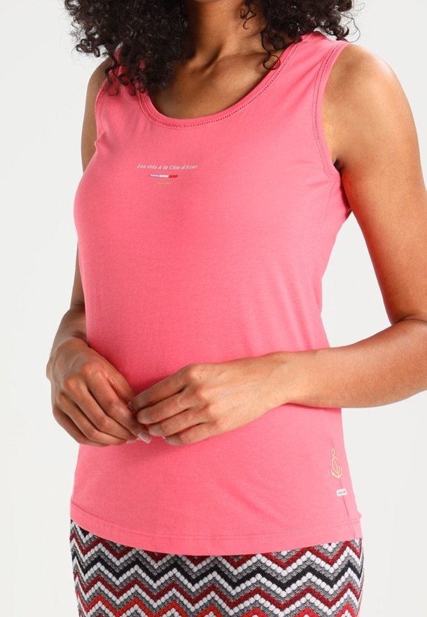 Femme portant un débardeur rose sans manches avec un petit texte blanc et une jupe à motif zigzag, les mains légèrement jointes devant.