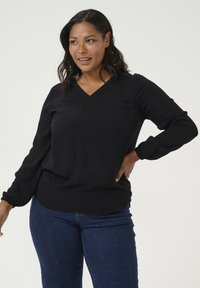 T-shirt noir à manches longues avec un décolleté en V, tissu texturé, poignets froncés et coupe décontractée, associé à un jean bleu foncé.