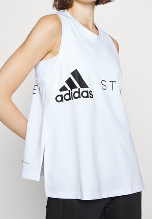 Frau trägt ein ärmelloses weißes Adidas Workout-Top mit schwarzem Logo und schwarze Leggings, steht mit den Händen hinter dem Rücken.