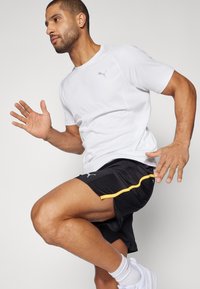 Camiseta deportiva blanca con logo gris, combinada con pantalones cortos negros con detalles amarillos. El modelo muestra una pose dinámica de carrera.