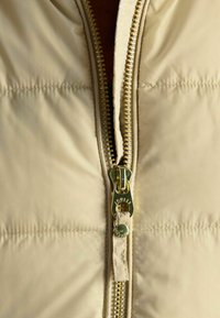 Bežová prešívaná bunda s kovovým zipsom. Tkanina je hladká s prešívanými sekciami, čo ukazuje minimalistický dizajn a značkový akcent.
